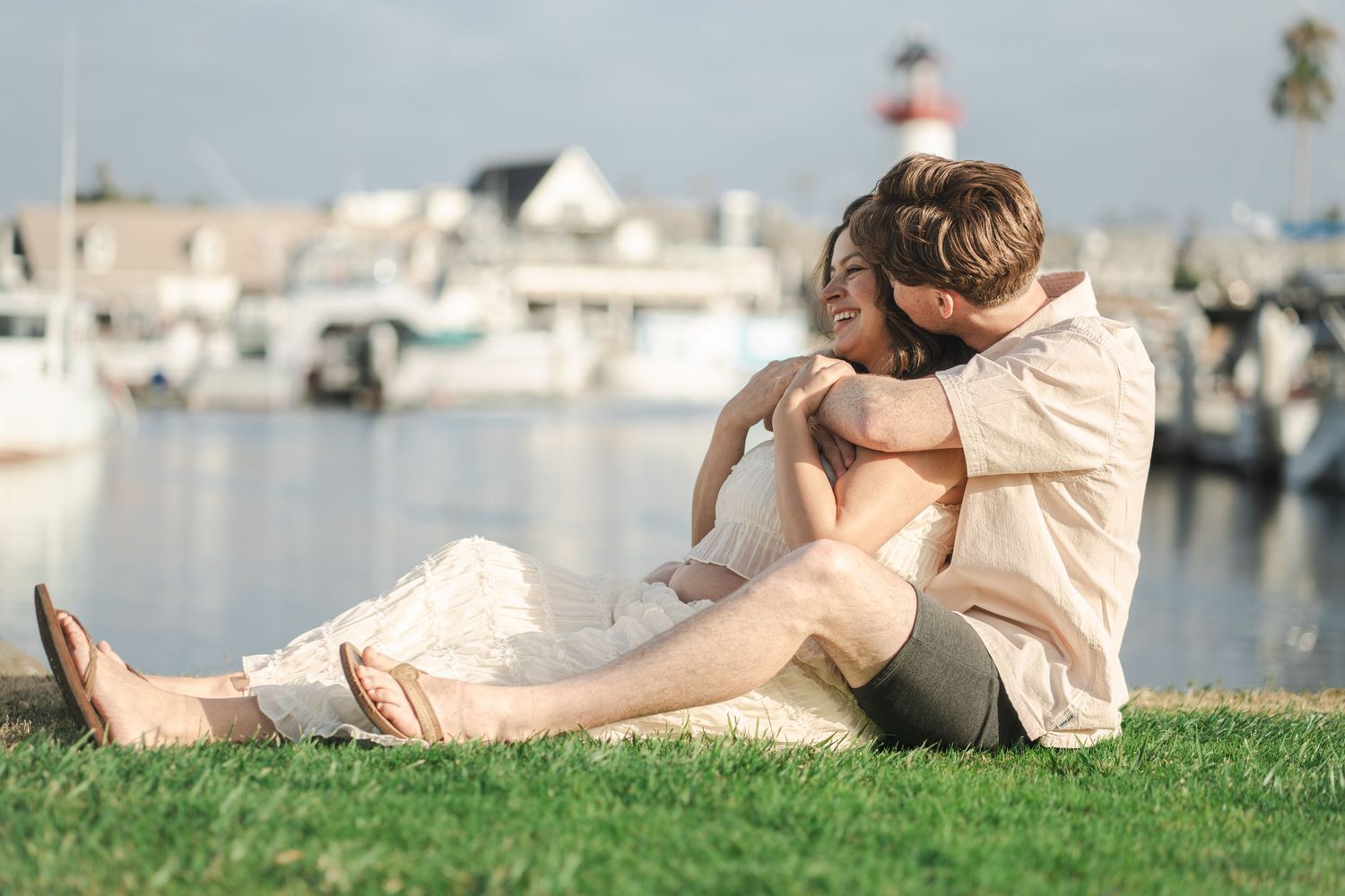 A couple sits embracing on green grass by a marina, sharing a romantic moment with boats and a lighthouse in the background. Oceanside, CA