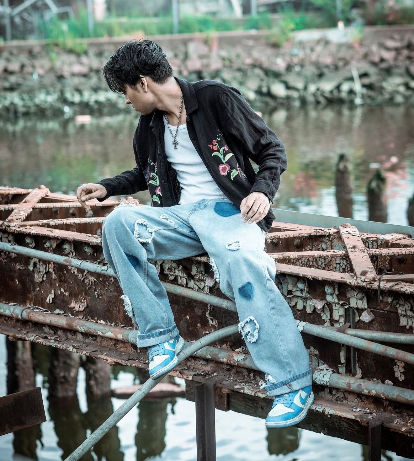 Young man in black floral jacket and ripped jeans sitting on rusty metal structure near water.