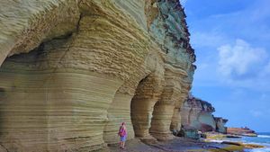 Person standing beside massive layered sandstone rock formations on a coastal cliff under a blue sky.