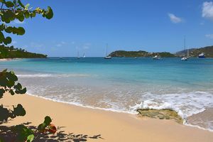 Turquoise Caribbean beach with white sand, sailboats anchored in calm blue waters, and lush green hills under a clear sky.