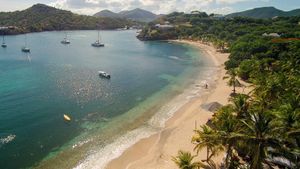 Aerial view of a tropical beach with turquoise water, sailboats, golden sand, and lush palm trees in a scenic bay.