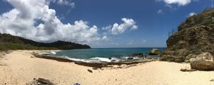 Panoramic view of a secluded tropical beach with turquoise waters, rocky cliffs, and lush green hills under a partly cloudy sky.