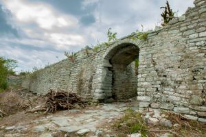 Ancient stone archway in ruins with overgrown vegetation and cobblestone path, historic medieval fortress wall.