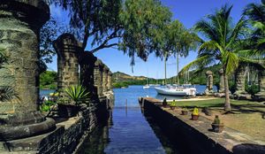 Historic stone ruins frame a serene harbor with sailboats, palm trees, and lush greenery in Antigua, Caribbean.