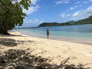 Person standing on a white sandy tropical beach with clear turquoise water, a pier, and lush green hills in the background.