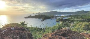 Panoramic sunset view of a tropical harbor with sailboats, lush green hills, and a coastal village in Antigua.
