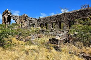 Ancient stone ruins with arched walls surrounded by dry grass and shrubs under a bright blue sky.
