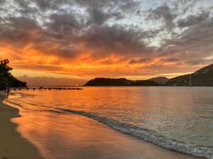 Stunning tropical beach sunset with vivid orange and red sky reflecting on calm ocean waters, sandy shore, and distant hills.