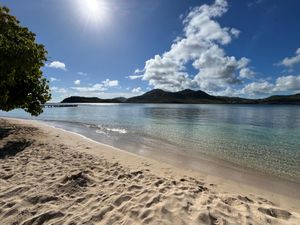 Pristine white sandy beach with crystal clear turquoise water and lush green hills in background.