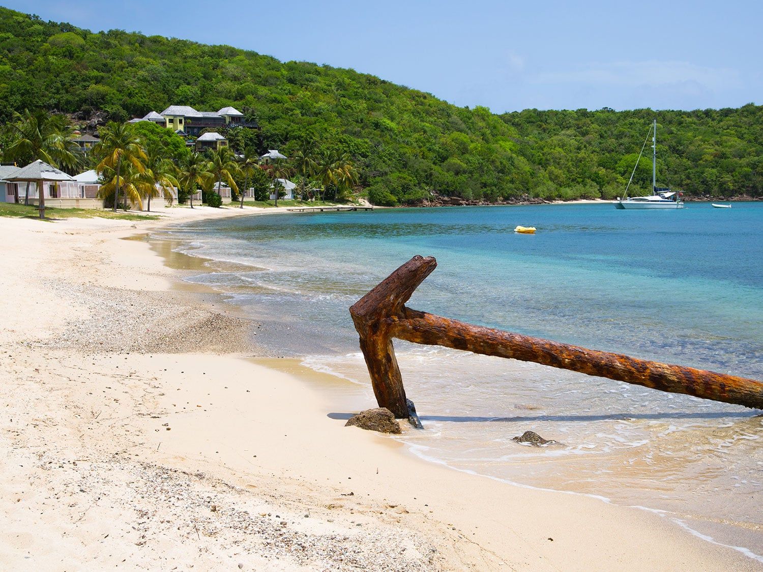 Rusty anchor on a tropical sandy beach with turquoise water, sailboat, and lush green hills in the background.