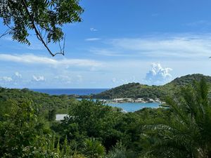 Lush tropical hillside overlooking a turquoise Caribbean bay surrounded by green trees under a bright blue sky.