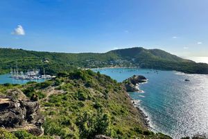 Aerial view of a lush tropical bay with sailboats, turquoise waters, and green hills under a bright blue sky.