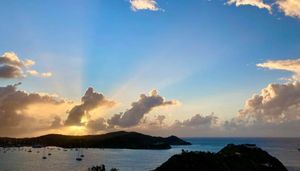 Stunning tropical sunset with golden rays breaking through clouds over a calm bay with boats and lush green islands.