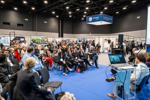 Large audience seated on blue chairs at an indoor conference expo hall during a professional presentation event.