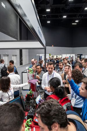 Crowded tech expo hall with attendees gathered around vendor booths displaying electronics and 3D printers at a convention center.