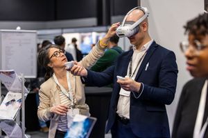Woman helping man try on VR headset at a tech conference exhibition booth with attendees in background.