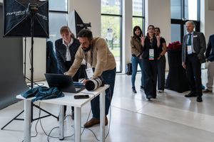 Professional photographer reviews images on laptop at indoor event setup with studio lighting and attendees in background.