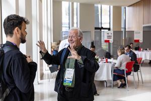 Two men engaged in conversation at a conference venue, with attendees seated at tables in the background.
