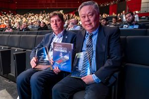 Two men seated in auditorium holding awards and Applied Photonics books at a professional conference.