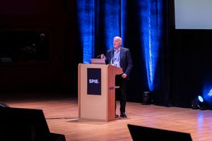 Speaker presenting at SPIE conference podium on stage in a large auditorium with blue lighting backdrop.