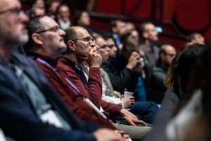 Audience members seated in rows at a conference, attentively listening to a presentation in a dimly lit venue.