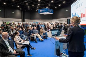 A presenter speaks at a packed conference expo hall with audience seated on blue chairs before a large presentation screen.