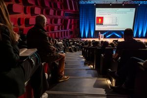 Audience members attend a large conference presentation with a speaker on stage and a bright projection screen.