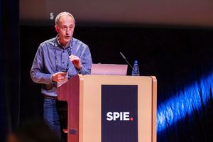 A speaker presents at an SPIE conference podium, wearing a blue shirt in a modern auditorium setting.