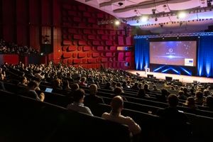 Large conference auditorium filled with attendees watching a presenter on a lit stage with projection screen.