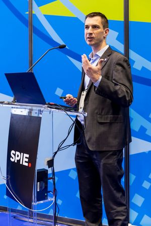 Male speaker gestures at SPIE conference podium with laptop during professional photonics industry presentation.