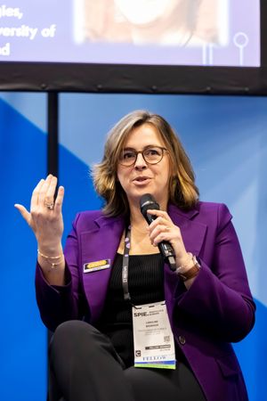 Woman in purple blazer speaks at SPIE conference, holding microphone and gesturing on stage.