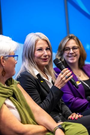 Three women sit on a panel stage, center speaker holds microphone, smiling, blue background at conference event.