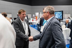 Two businessmen shaking hands at a professional trade show or conference event in a modern exhibition hall.