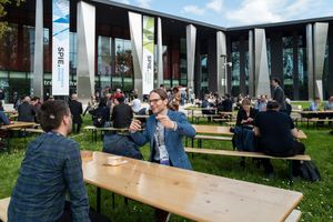 People networking outdoors at a tech conference, seated at wooden picnic tables on green grass near a modern building.