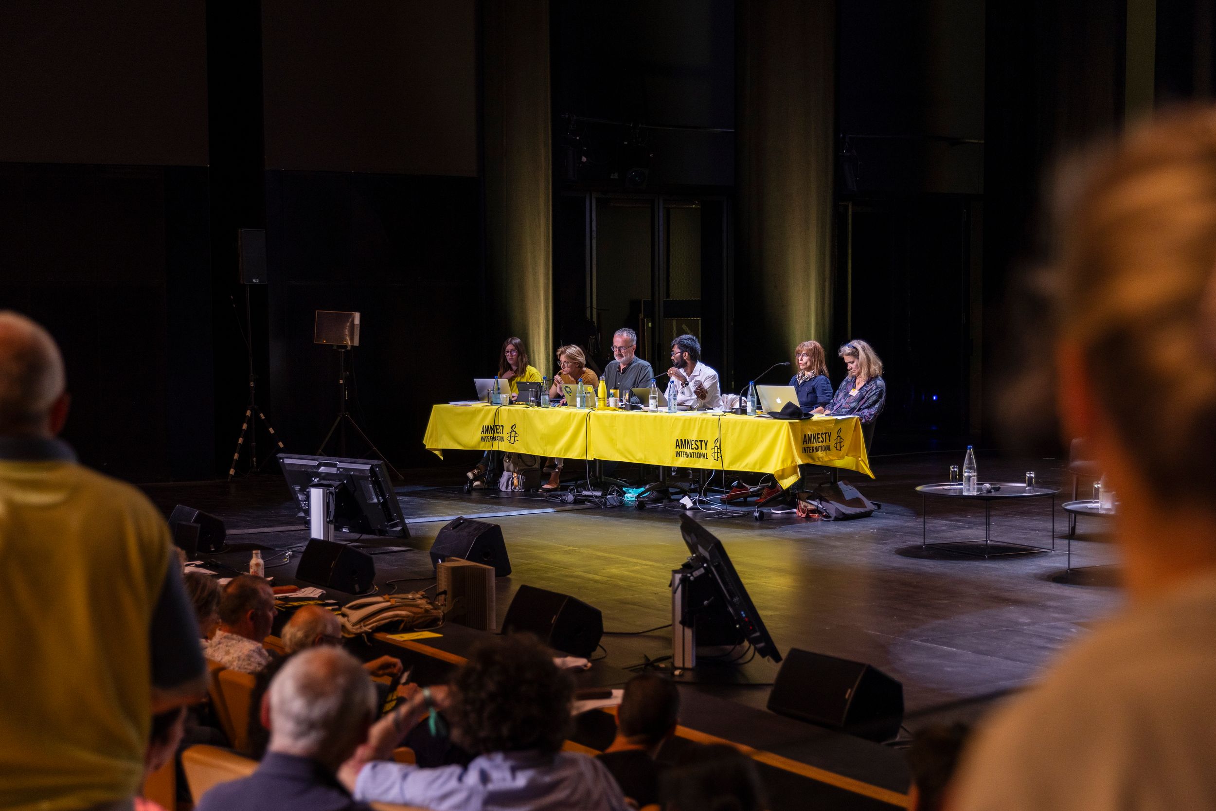 Panel discussion on stage with yellow tablecloth, speakers seated before an audience in a darkened theater.