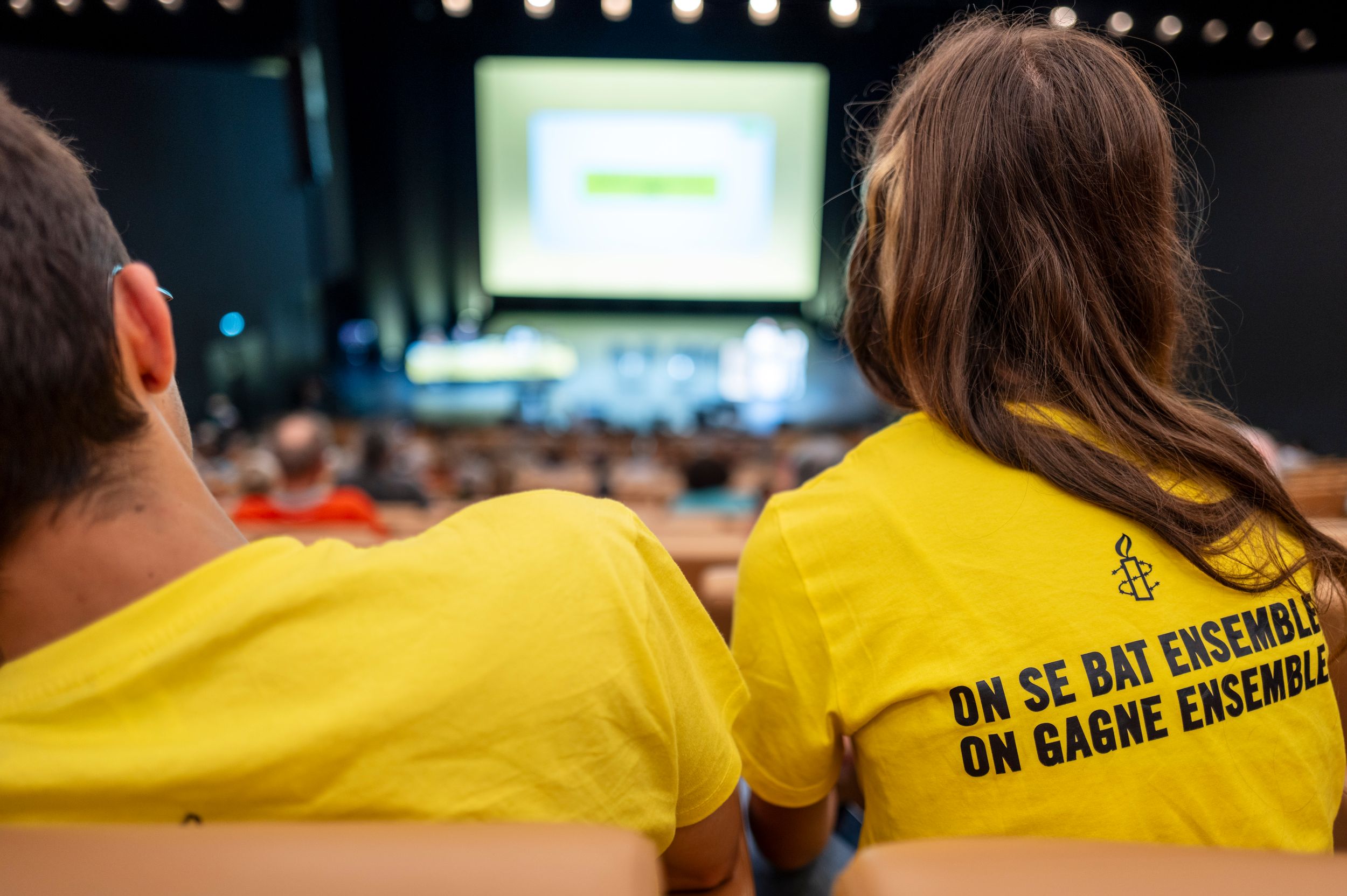 Two people in yellow Amnesty International t-shirts attend a conference, with text reading On Se Bat Ensemble On Gagne Ensemble.
