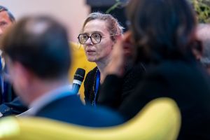 Woman with glasses speaks into microphone at professional conference panel discussion event.