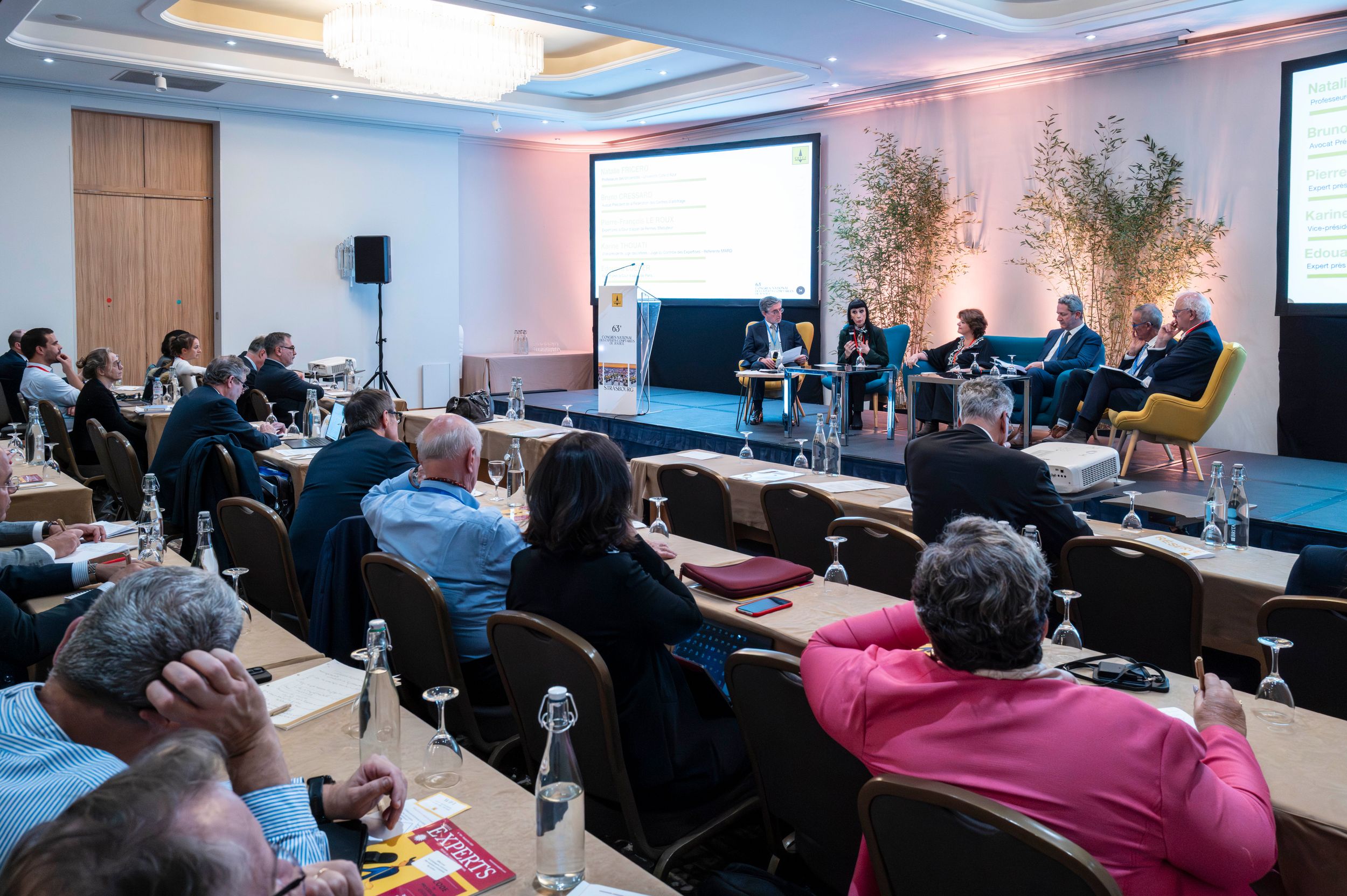 Business conference panel discussion with audience seated at tables facing speakers on a stage with a presentation screen.