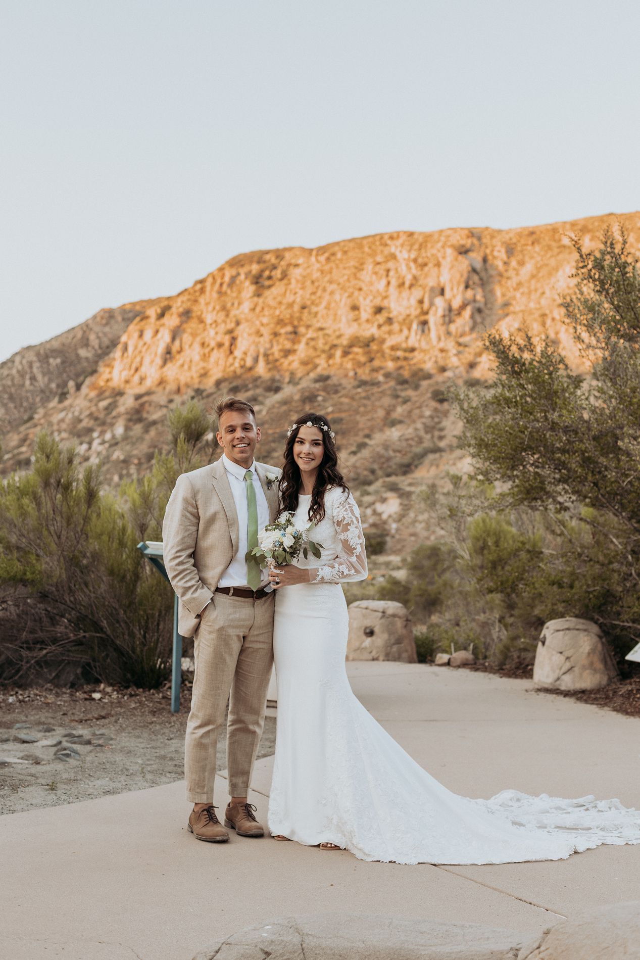 A smiling bride and groom pose together in front of a rocky desert mountain landscape after their outdoor wedding ceremony.