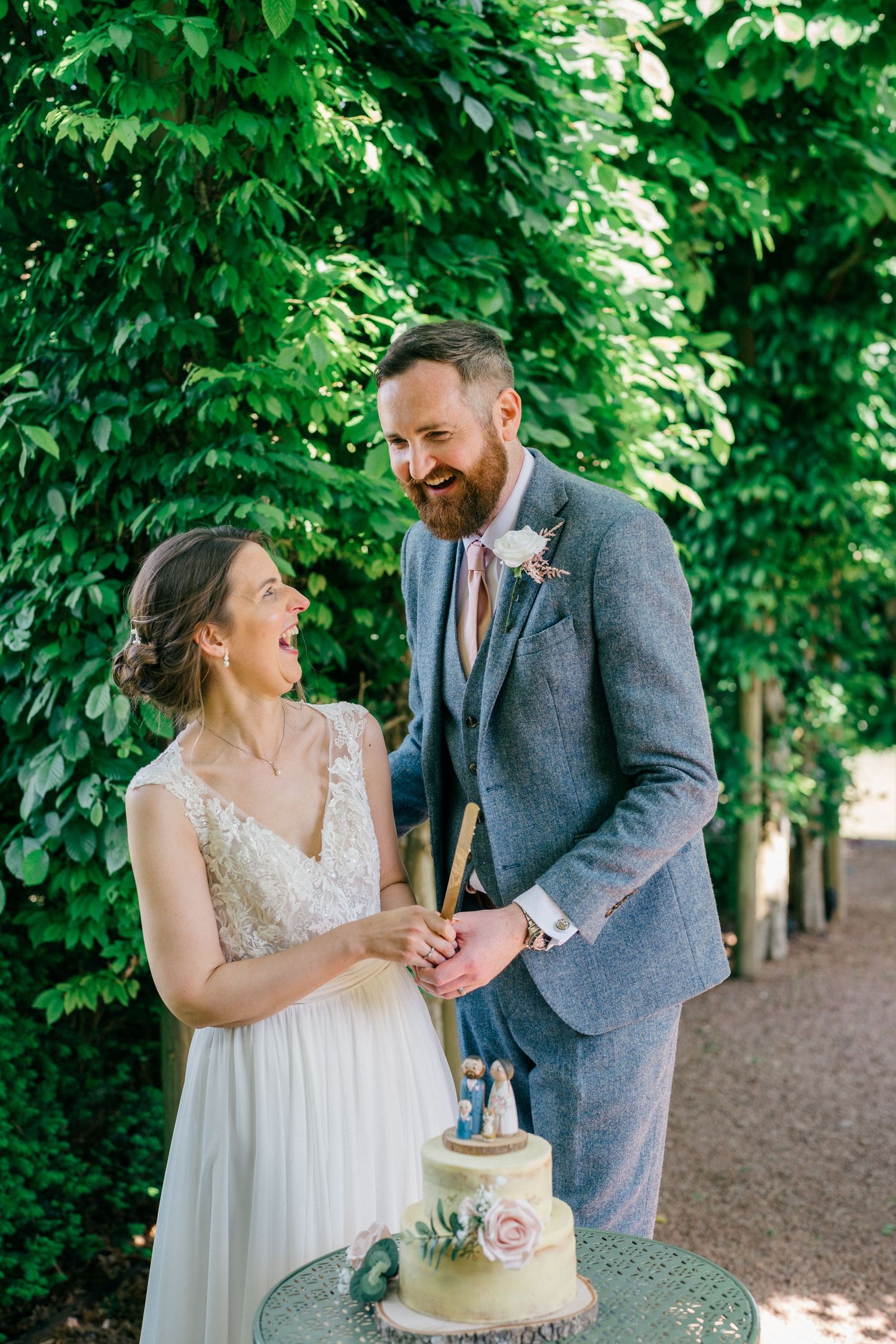 Bride and groom laughing while cutting their wedding cake outdoors, surrounded by lush green hedges.
