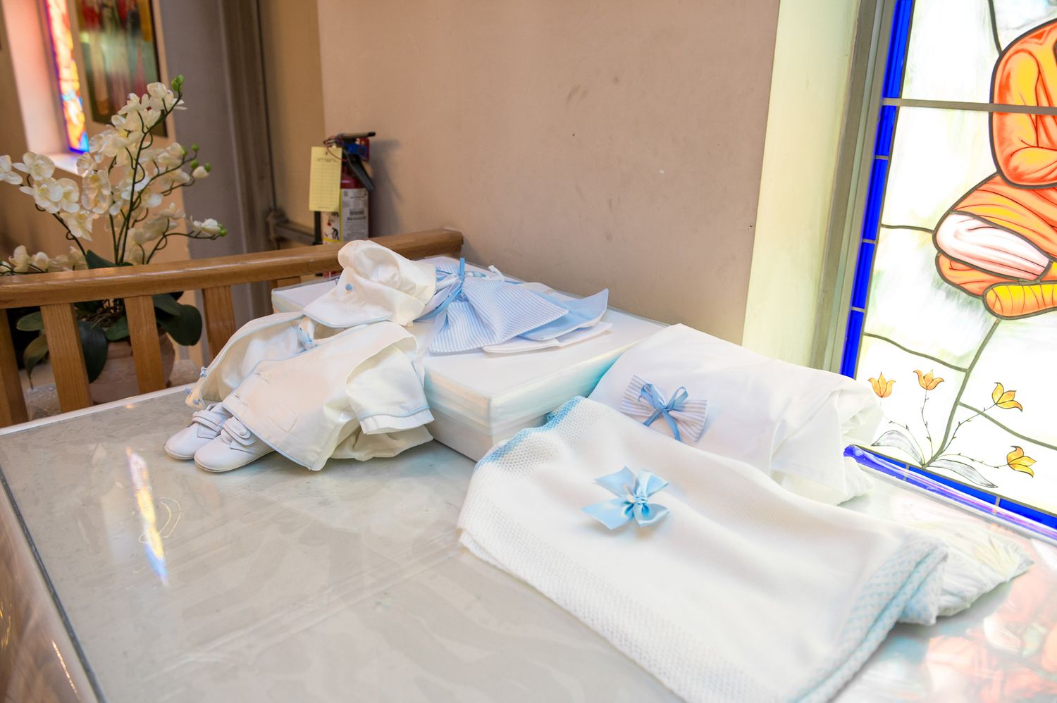White baptism outfit with blue bow accents and baby shoes laid out on a table near a stained glass window.