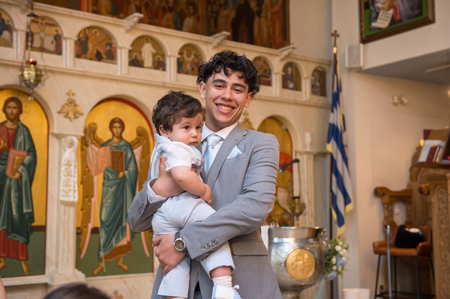 Young man in gray suit holding a baby dressed in white during a Greek Orthodox baptism ceremony.