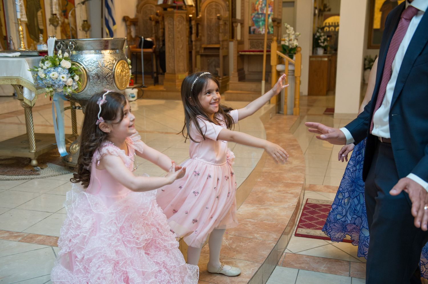 Two young girls in pink dresses dancing joyfully inside a decorated Greek Orthodox church.