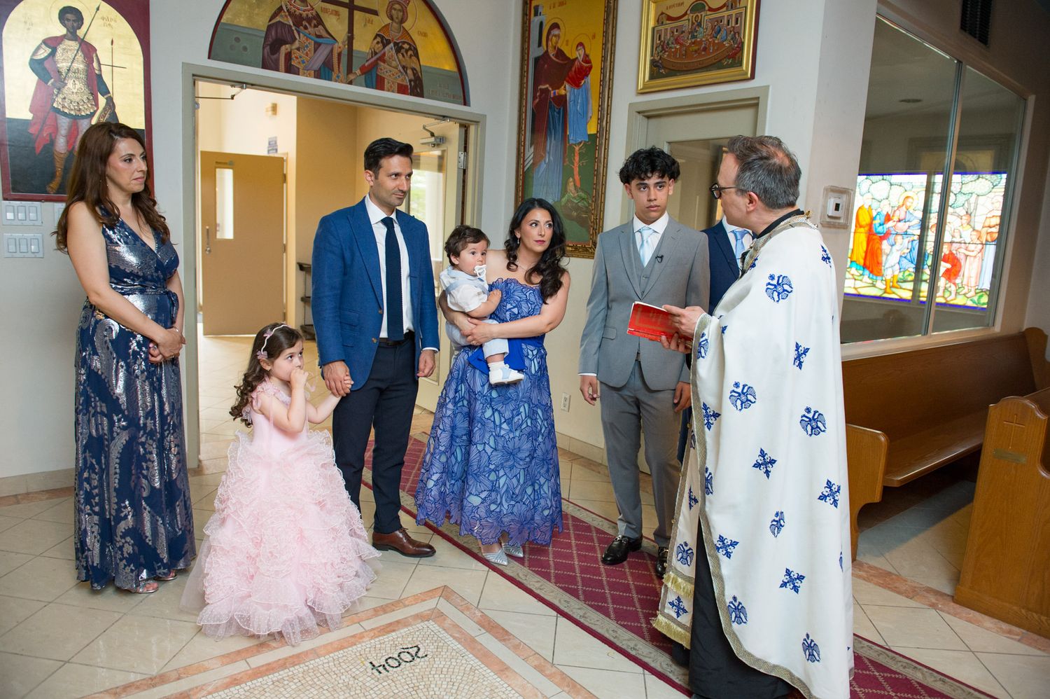 Family gathering with priest in ornate vestments during a Greek Orthodox baptism church ceremony.