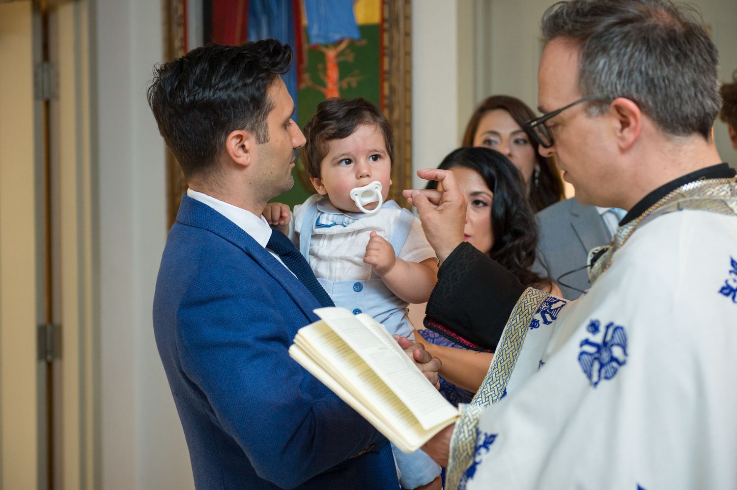 Greek Orthodox priest reading scripture while parents present their baby during a baptism ceremony.