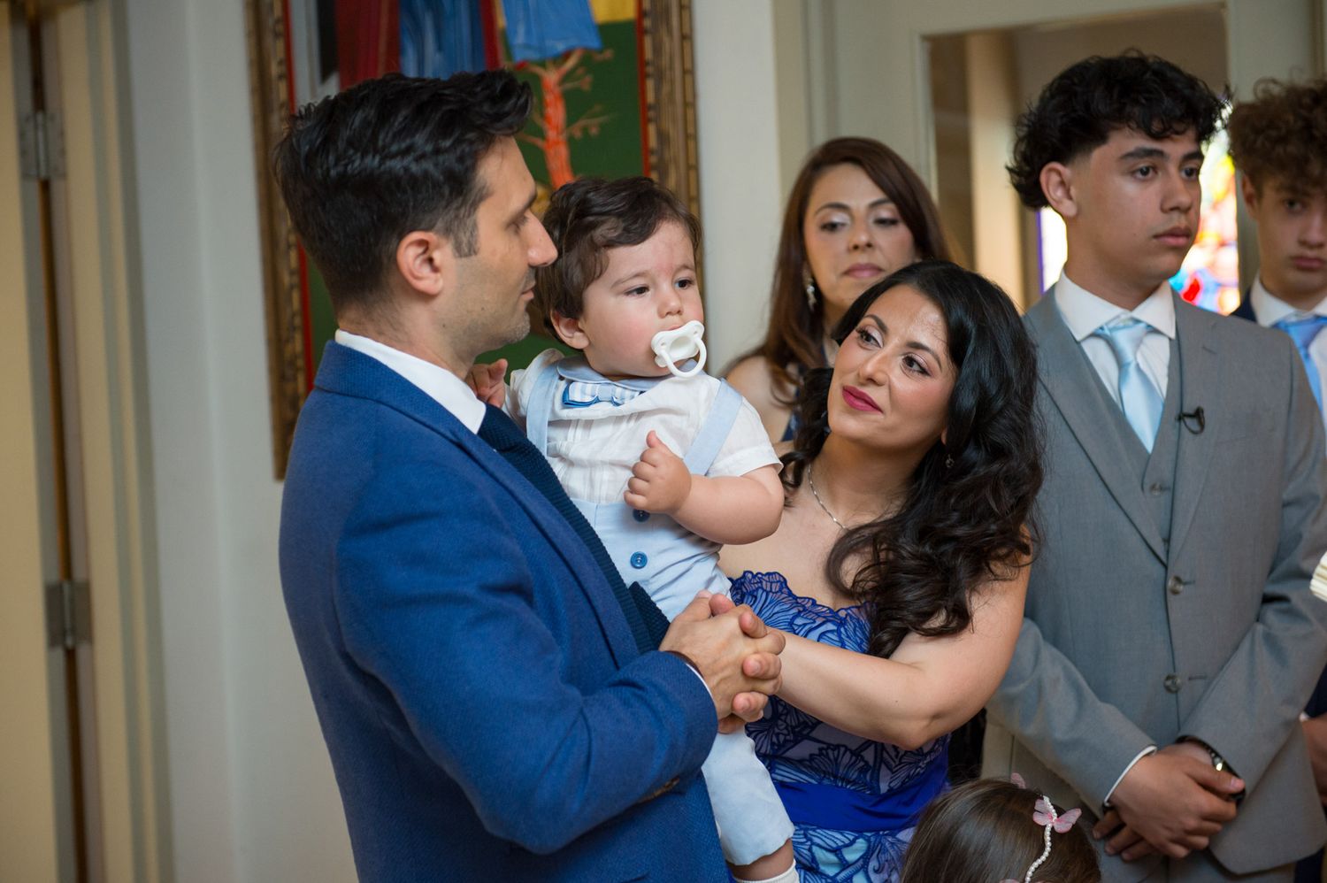 Parents in formal attire lovingly holding their baby boy during a Greek Orthodox baptism ceremony.