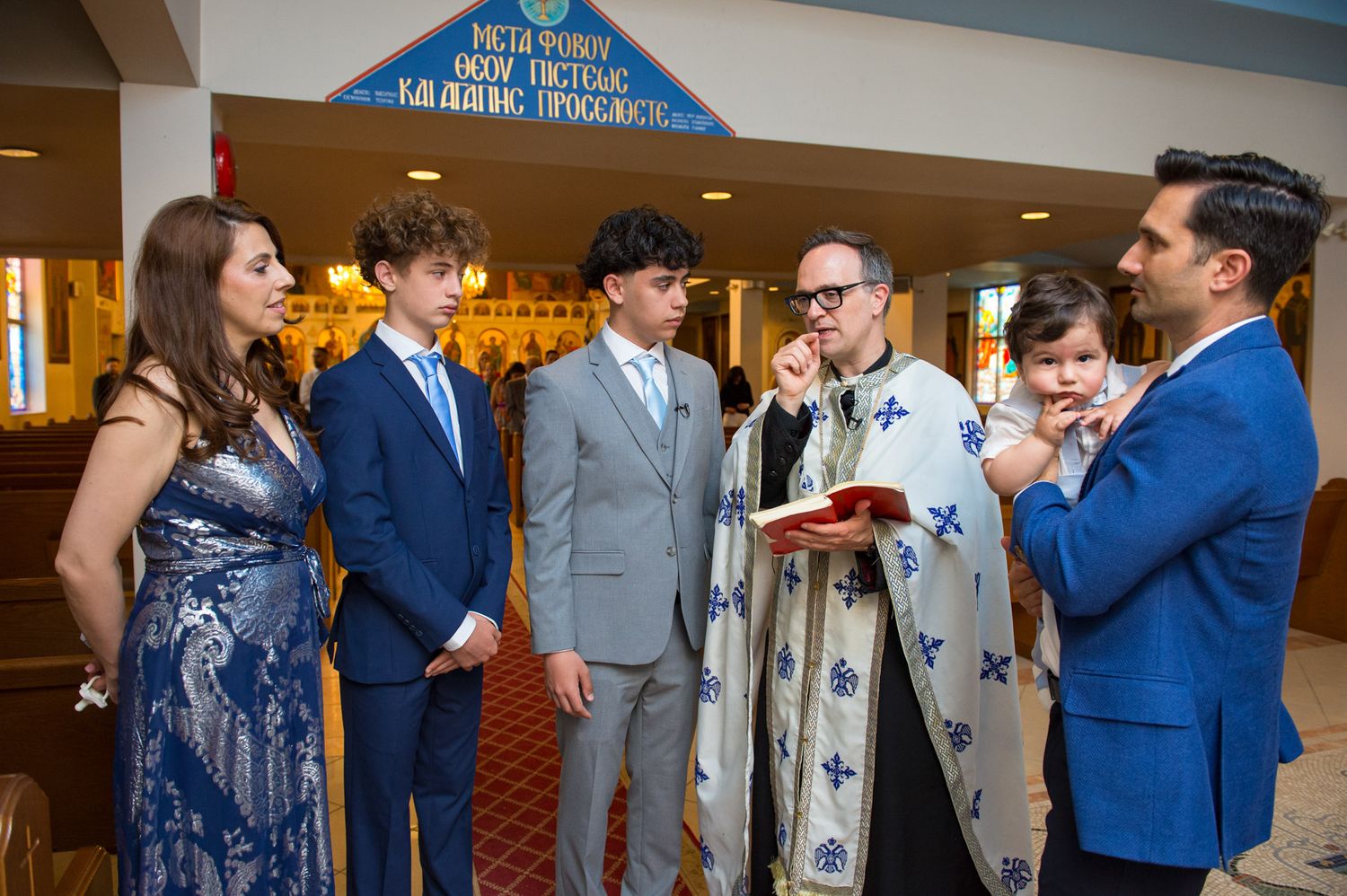 Greek Orthodox priest reading from a book with family members gathered during a baptism ceremony.