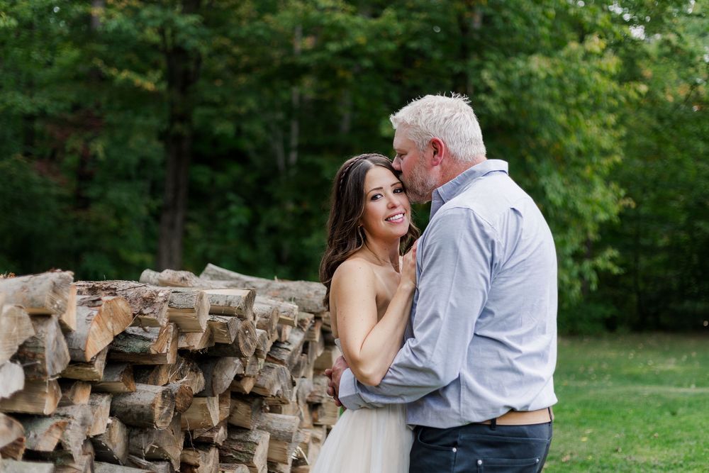 Bride and groom snuggle together near a wood pile during their backyard summer wedding for natural wedding photos near woodstock, ontario.