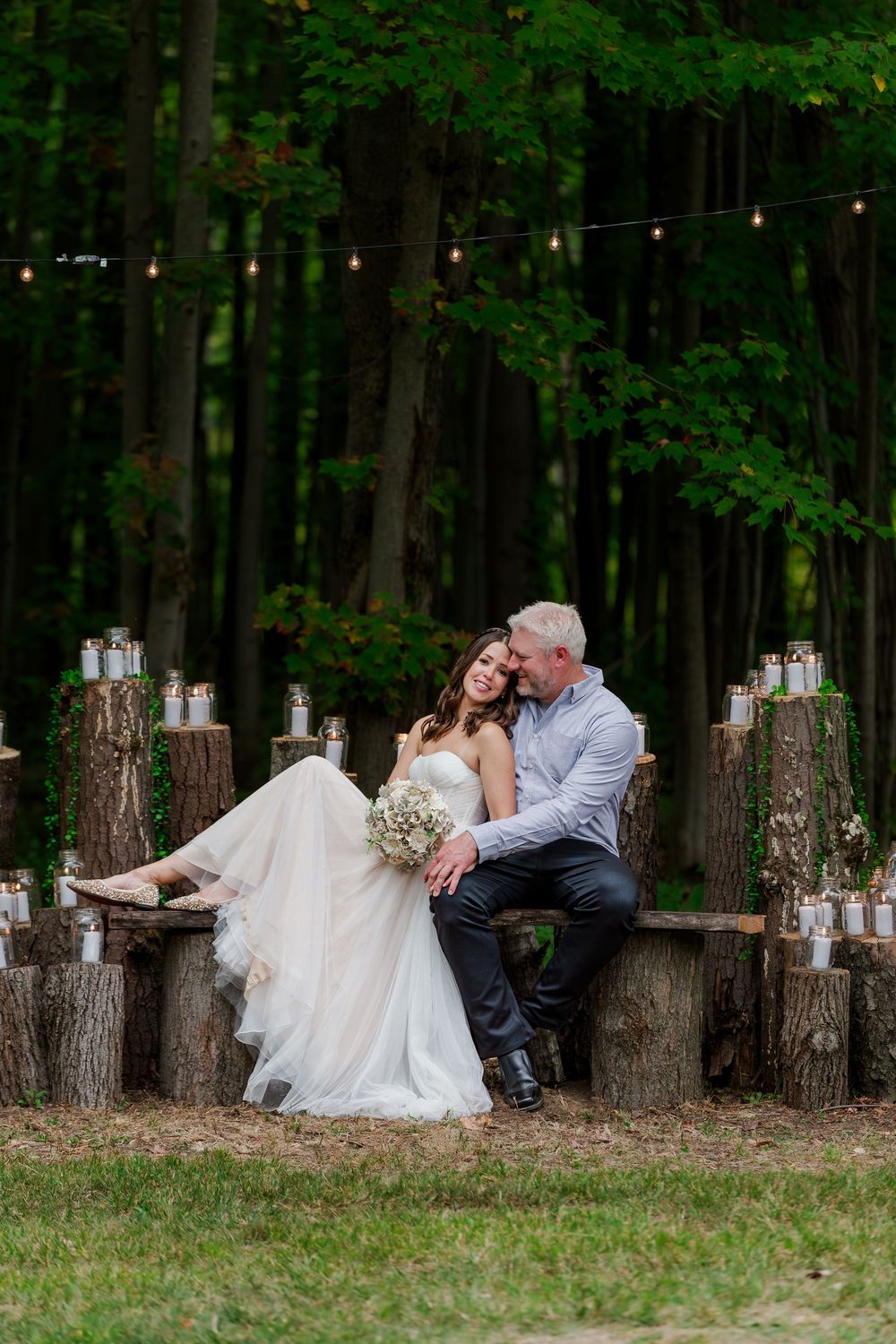 Bride and groom snuggle together on wood log bench surrounded by candles in their backyard summer wedding near woodstock, ontario.