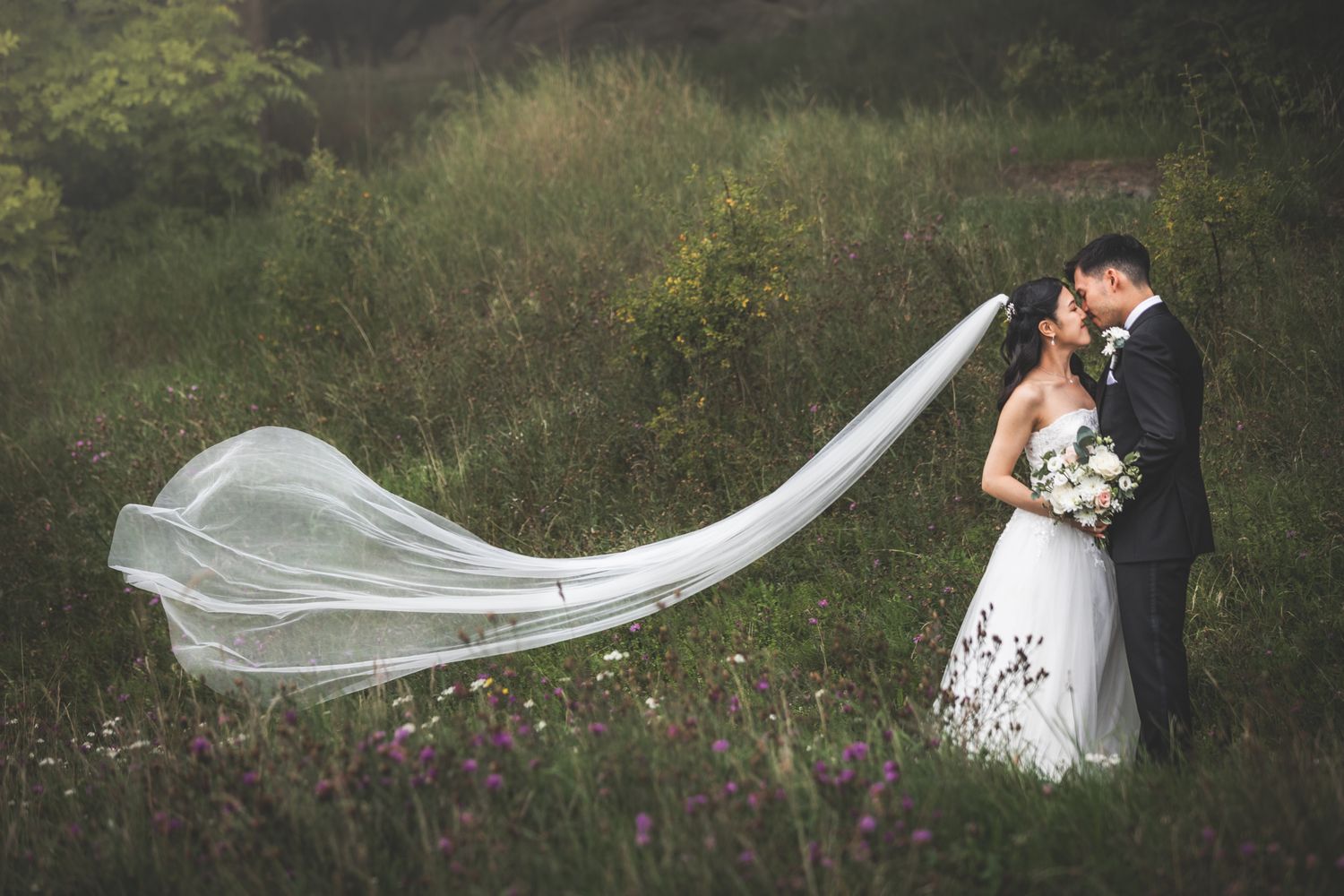 A flowing white wedding veil catches the breeze in a romantic outdoor garden setting with purple wildflowers.